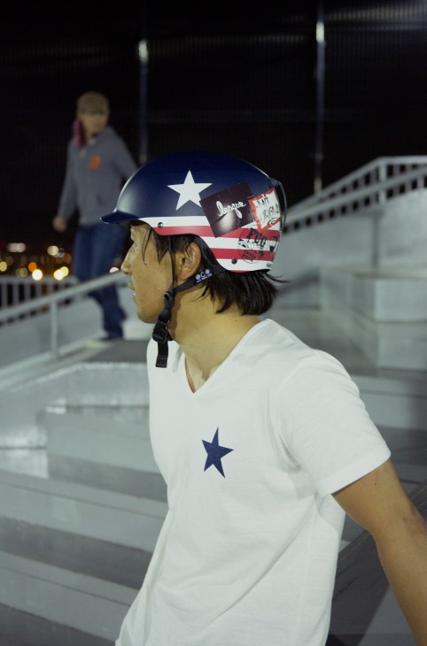 Student Hitoshi Watanabe wearing a patriotic helmet and white shirt with star design on a rooftop skate park at night.