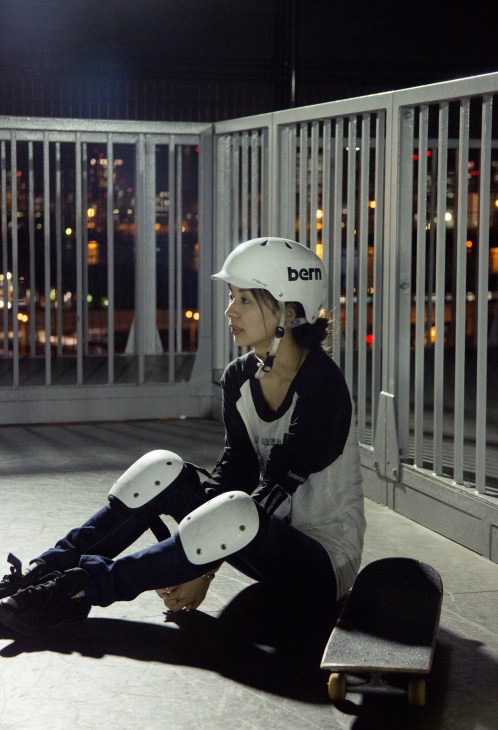 Person wearing a white bern helmet and knee pads sits on a skateboard at night on a rooftop with metal railings and city lights visible in the background.