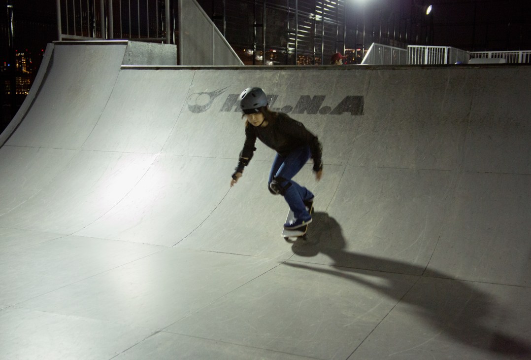 Young skateboarder performing a trick on an illuminated concrete ramp at night with city buildings in background.