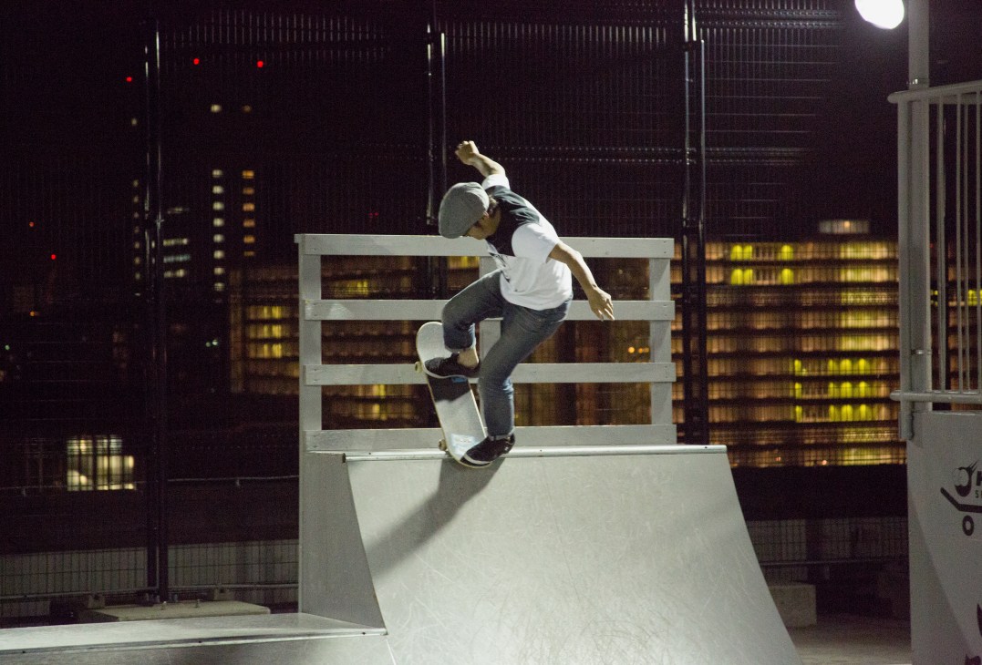 Skateboarding instructor Keita Ikenoue performs a trick on a white ramp against Tokyo's illuminated cityscape at night.