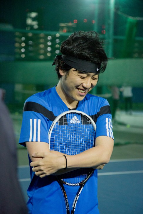 Tennis player in blue Adidas outfit holding racket, smiling during evening practice on illuminated court