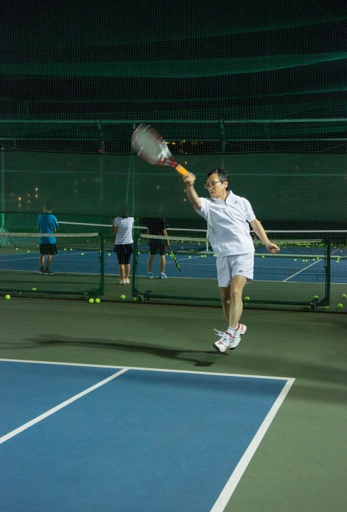 Tennis player serving on lit outdoor court with other players in background