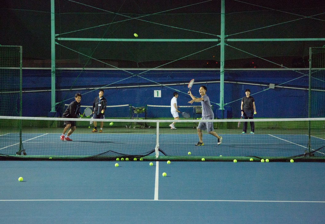 Tennis players practicing on a lit indoor court with green netting and blue court surface during evening lesson.