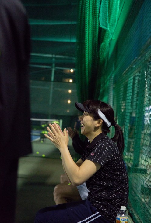 Woman in cap and polka-dot shirt clapping from sidelines of indoor tennis court.