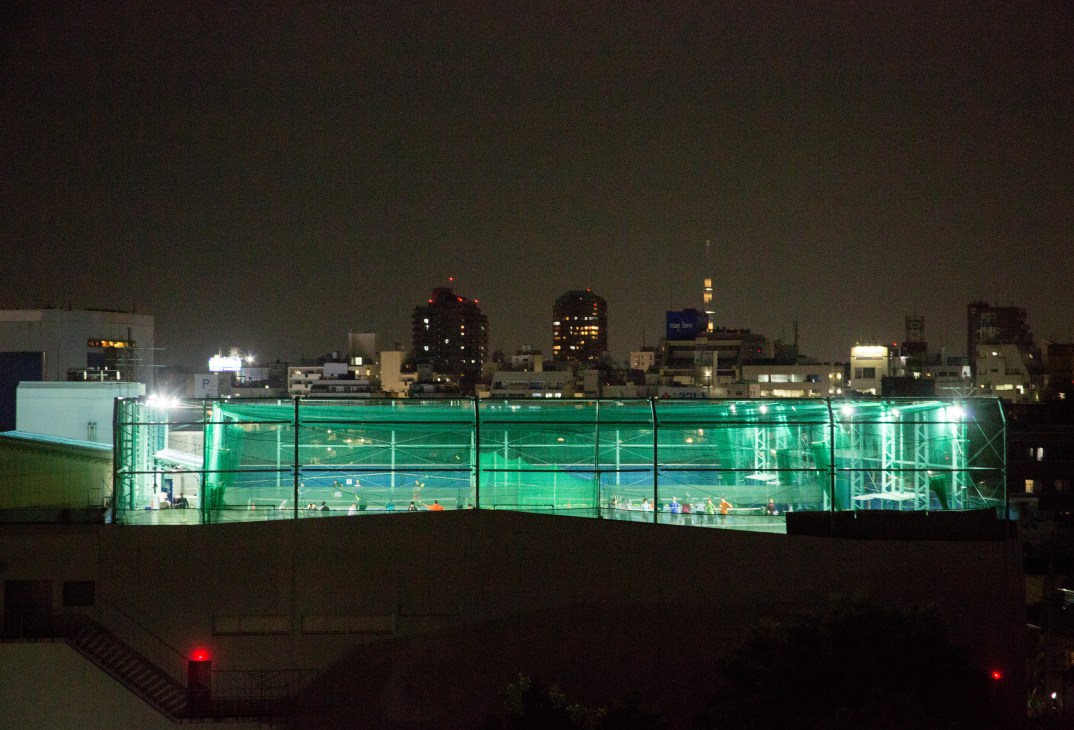 Illuminated rooftop tennis courts at night overlooking Tokyo's lit cityscape.