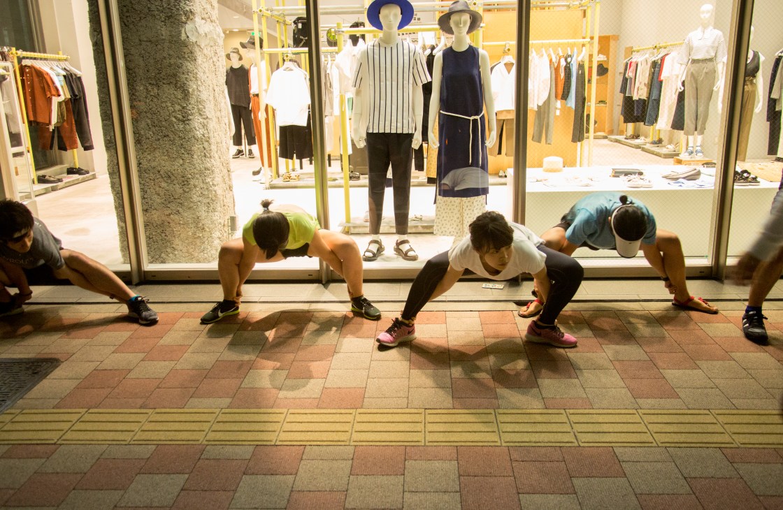Five runners stretching in forward bends outside a fashion store with mannequins visible in the window display.