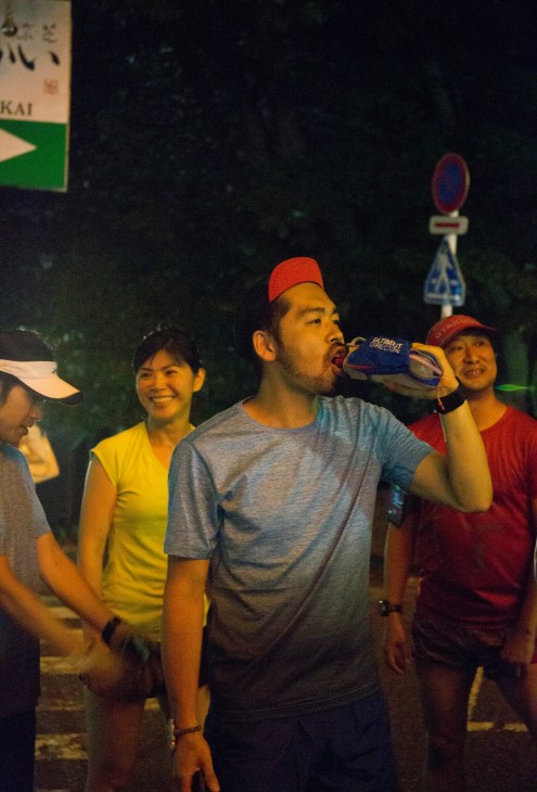 A man in a red cap drinks from a bottle while standing with smiling friends during a nighttime gathering in Tokyo.