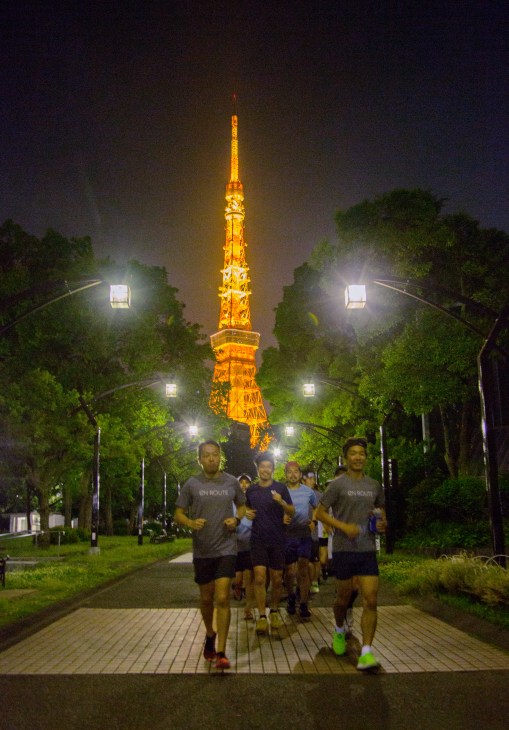 Group of runners jogging down a tree-lined path at night with Tokyo Tower illuminated in the background.