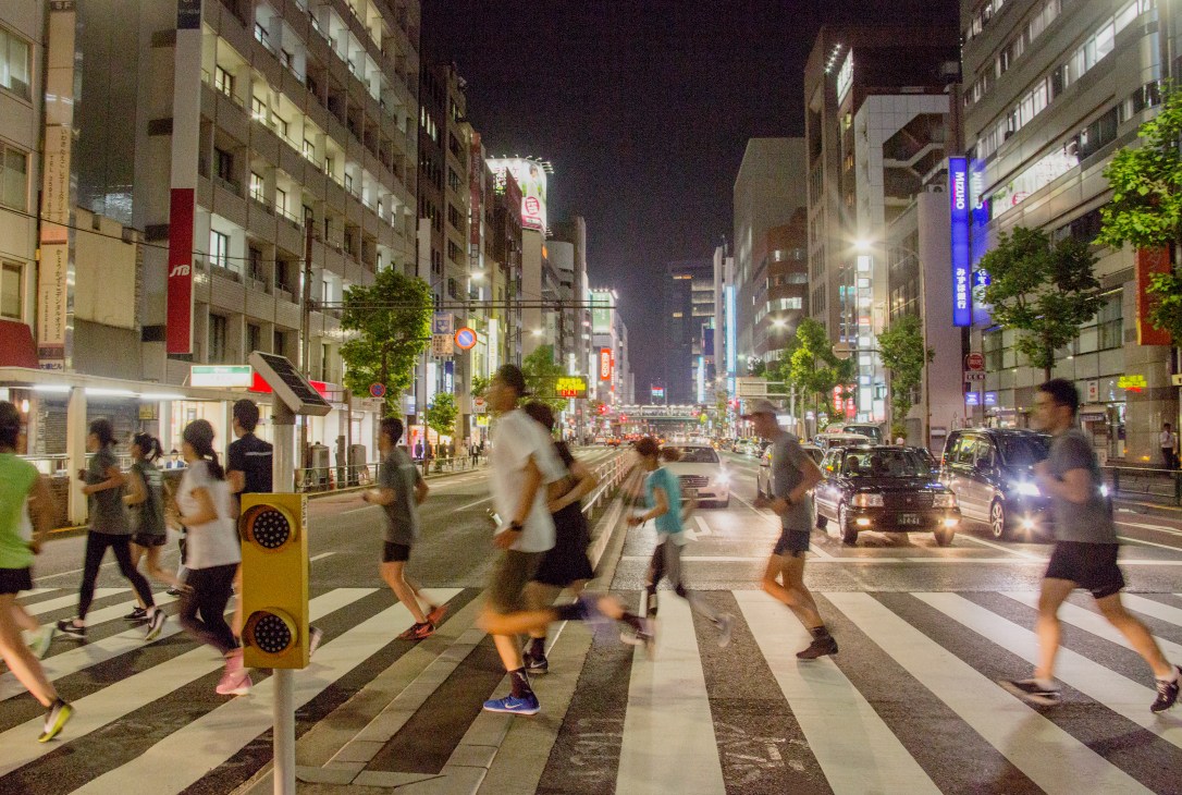 Runners crossing a zebra crossing in Tokyo's Shibuya district at night, surrounded by illuminated buildings and storefronts.