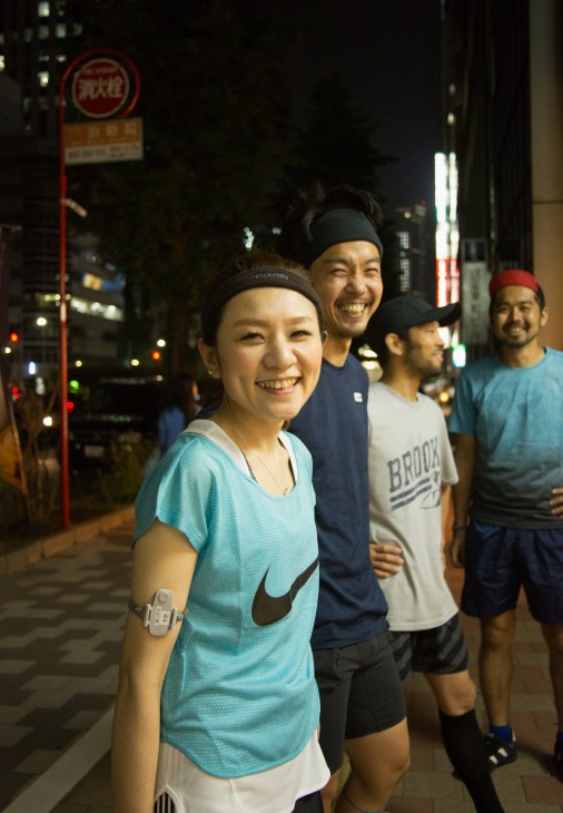 Four smiling runners in athletic wear stand together on a Tokyo street at night.