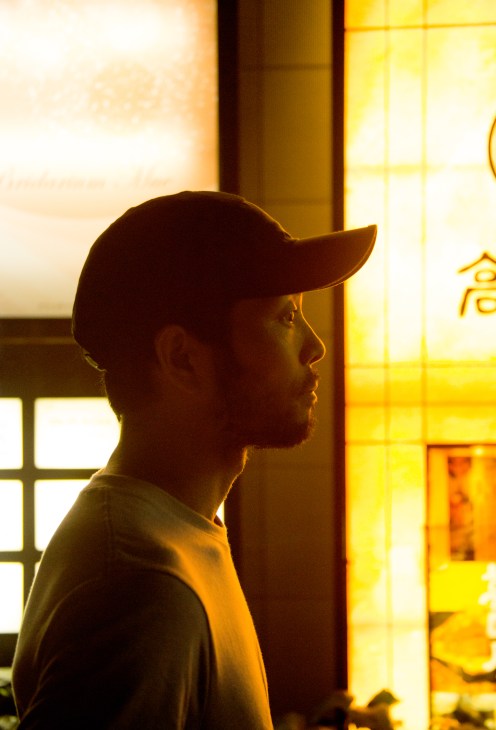 Man wearing a cap in profile against illuminated yellow storefront windows at night.