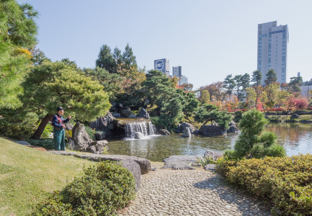 A person stands by a waterfall in a Japanese garden with autumn foliage and modern buildings in the background.