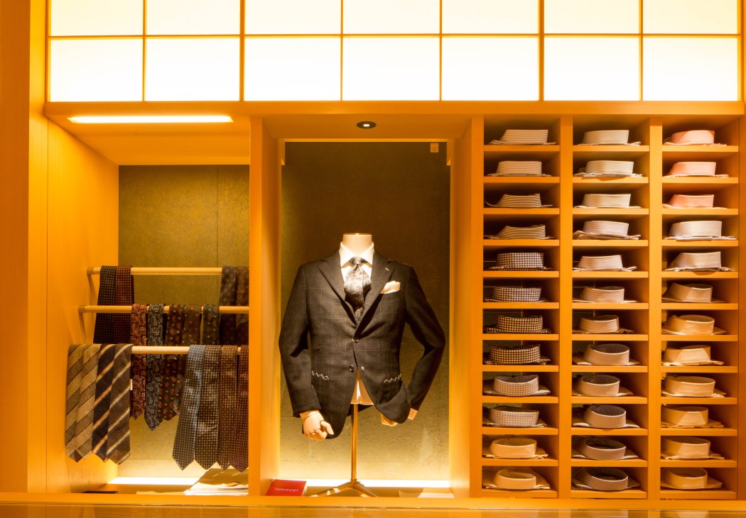 Elegant retail display with orange shelving showing menswear suit on mannequin and stacked hats.