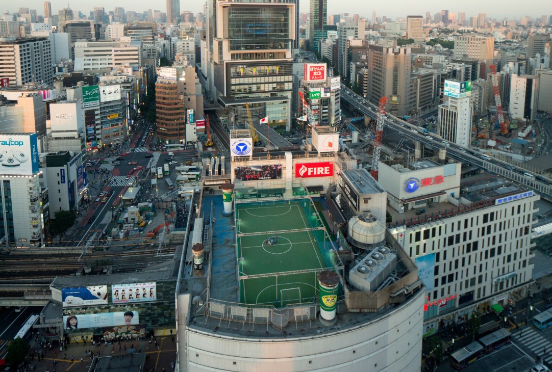 Aerial view of Tokyo's Shibuya district at dusk showing a rooftop futsal field with green playing surface surrounded by tall buildings and city streets below.