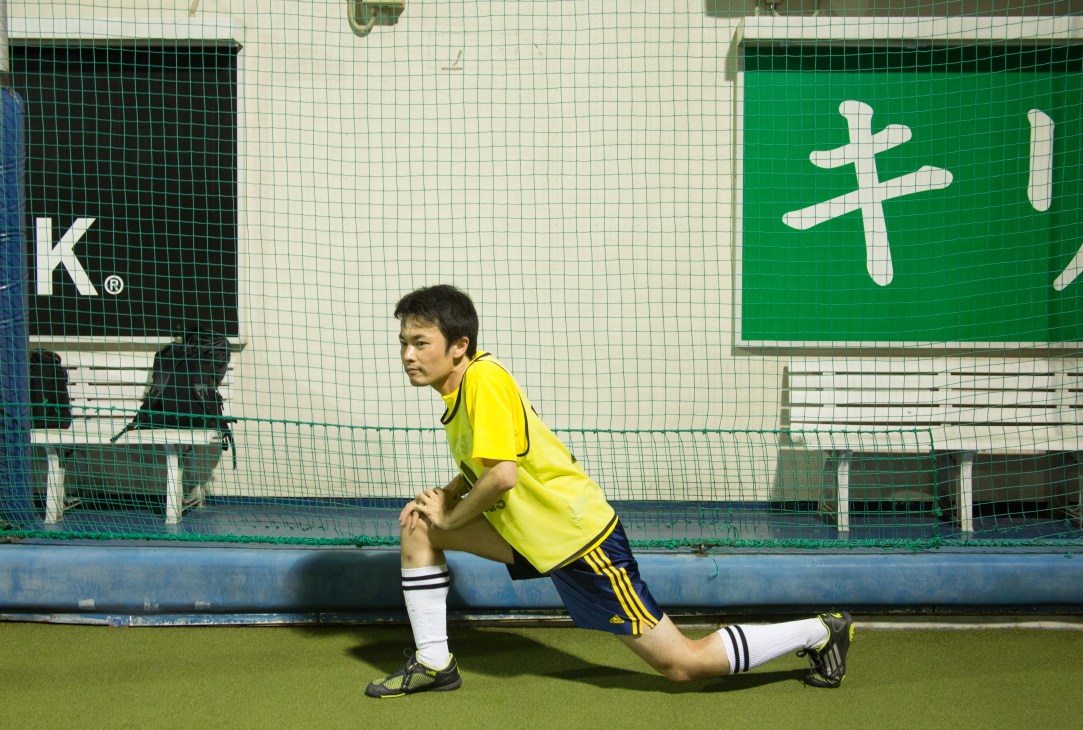 Young man in yellow sports shirt stretching on artificial turf with branded panels behind him.