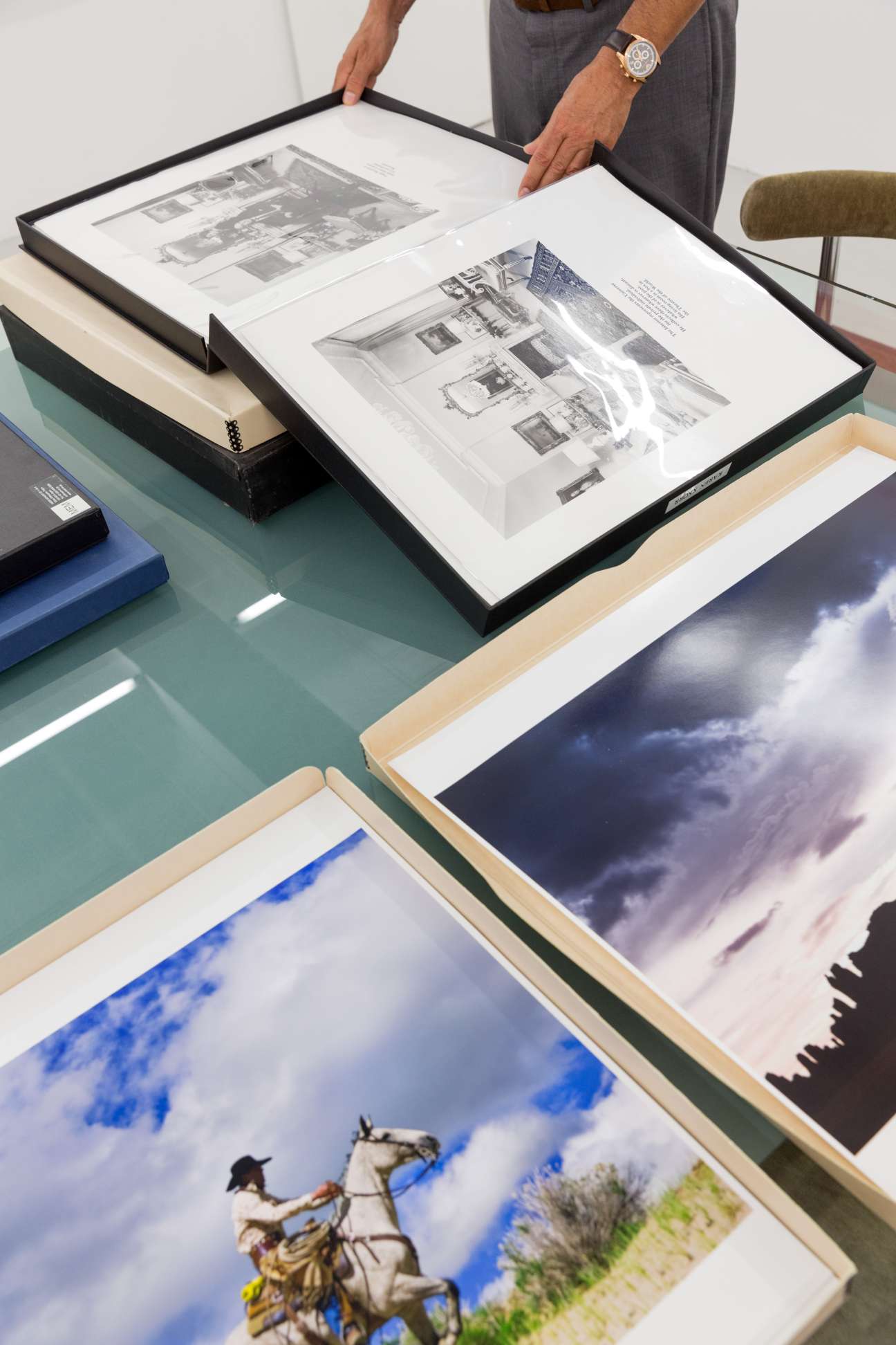 Person examining framed photographs and architectural drawings in portfolio boxes on a table.