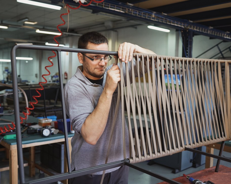 Craftsman weaving fabric by hand for outdoor furniture at a factory workstation.