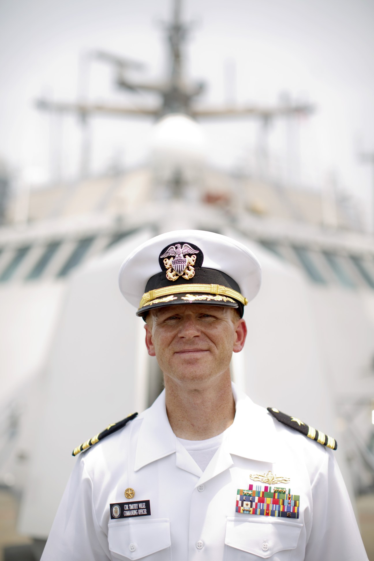 Commander Timothy Wilke in white Navy dress uniform with captain's hat stands on USS Freedom.