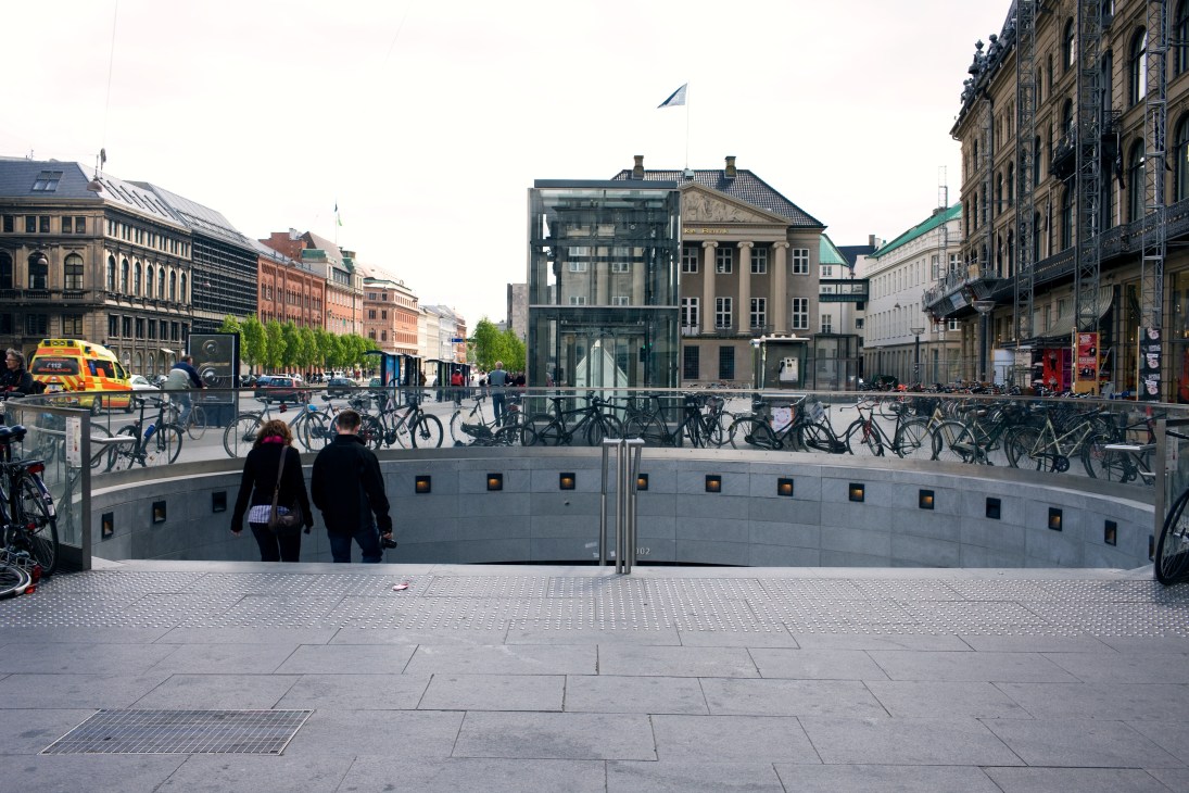 Subway entrance near Kongens Nytorv with cyclists and pedestrians in Copenhagen's urban plaza.
