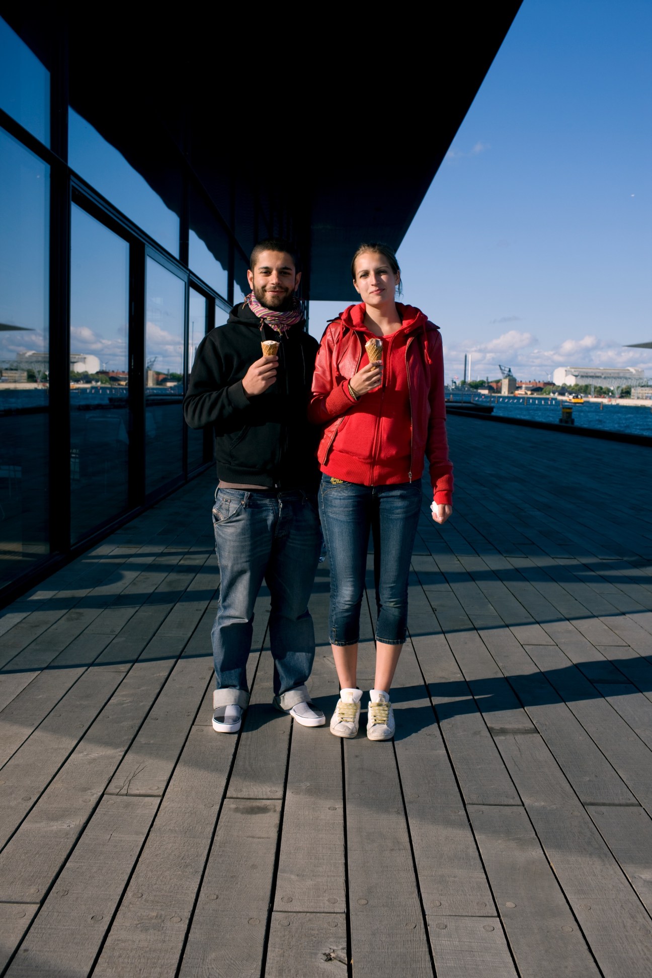 Two young people holding ice creams on a waterfront deck with Copenhagen harbor and buildings in the background.
