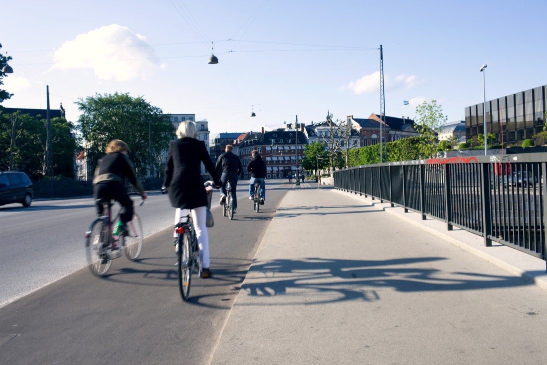 Multiple cyclists riding on a sunny bridge with modern railings and historic Copenhagen buildings in the background.