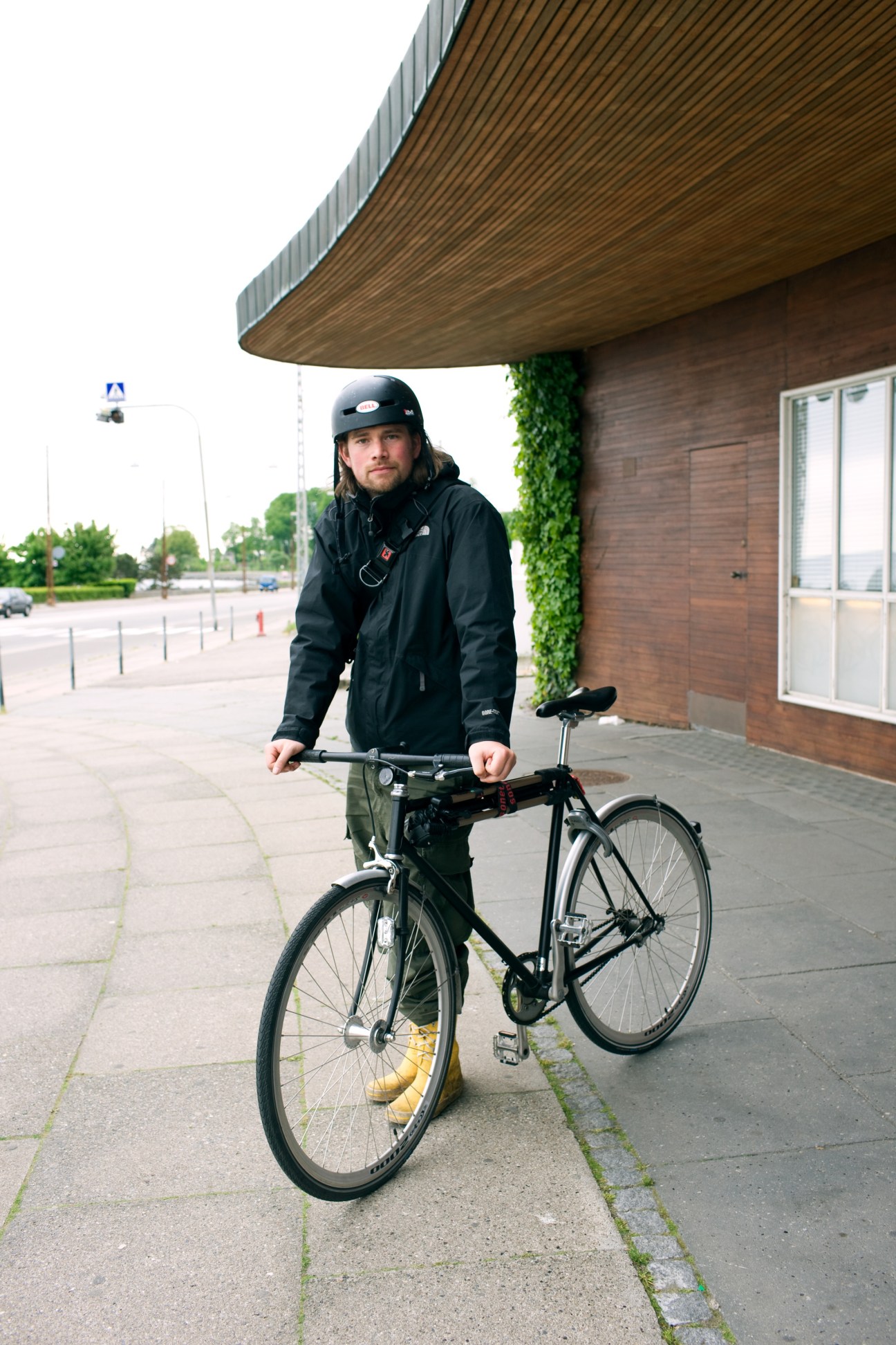 Man in black jacket and helmet standing with fixed-gear bicycle under modern wooden architectural overhang