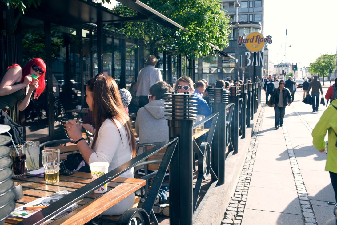 Outdoor café seating along a sunny Copenhagen street with pedestrians and a Hard Rock Café sign visible.