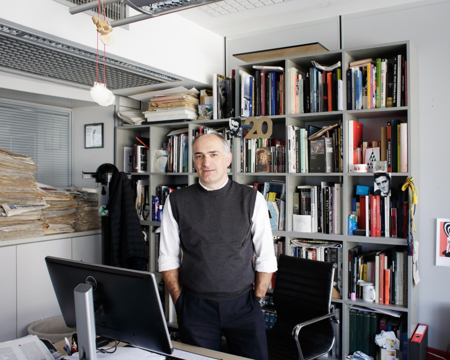Art director Angelo Rinaldi stands in his office in front of floor-to-ceiling shelves filled with books and design materials.
