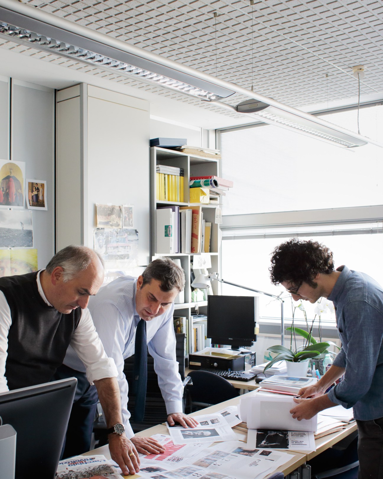 Three men examining layout designs and mock-ups on a work table in a bright office.