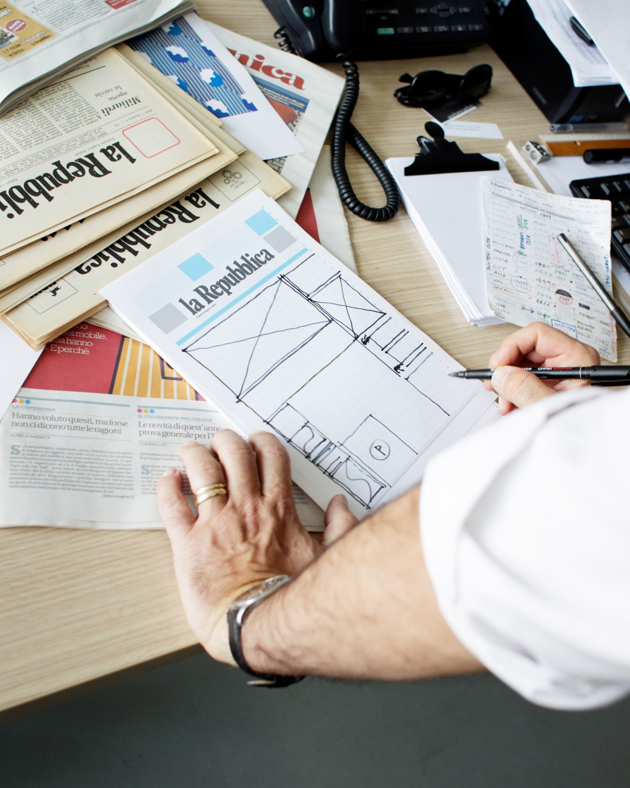 Art director Angelo Rinaldi sketches a layout on a front-page mock-up at his desk surrounded by La Repubblica newspapers and design materials.