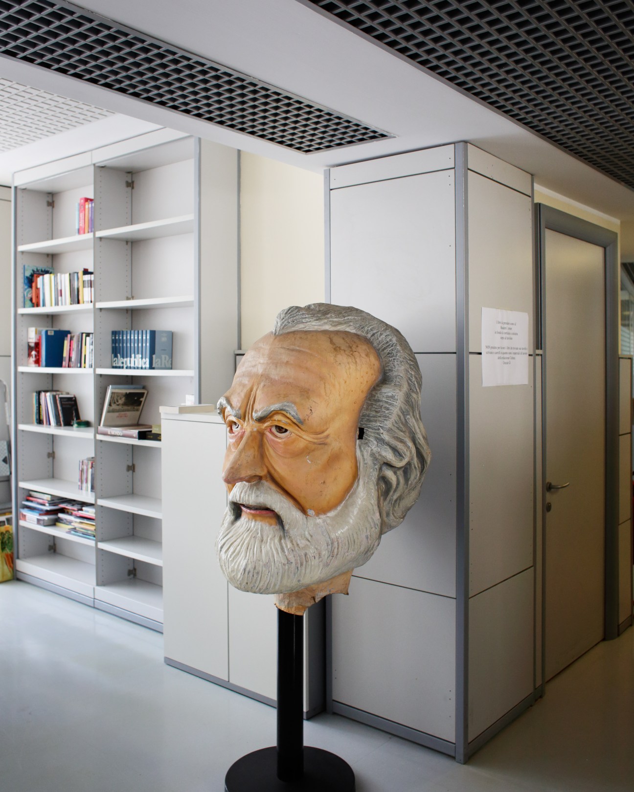 Wooden bust sculpture of bearded man with glasses on pedestal in modern office with white shelving.