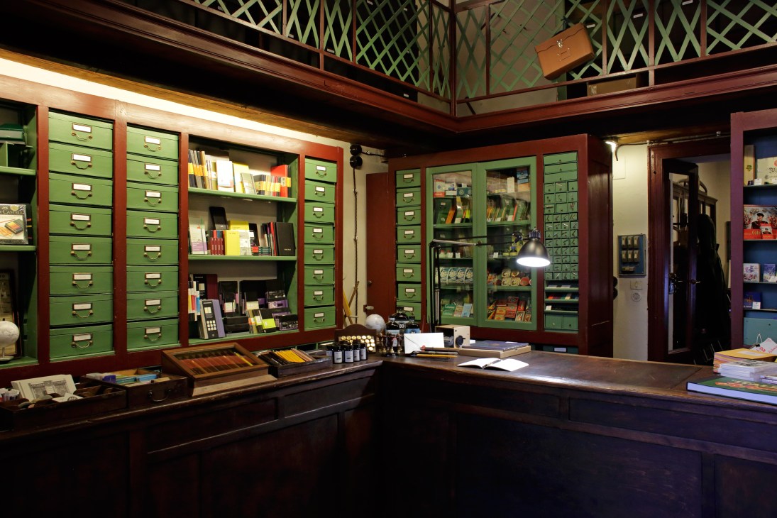 Interior of Fratelli Bonvini stationery shop with pea-green drawers, dark wood counter, and shelving displaying pens, papers and stationery supplies.