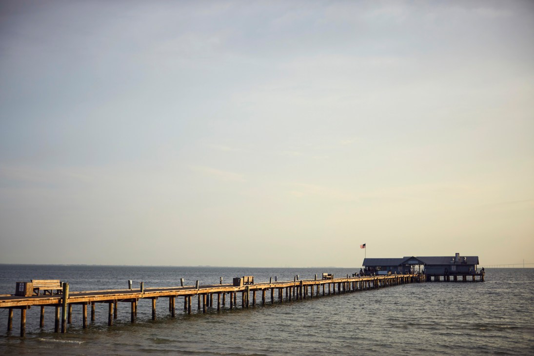 A long wooden pier extending into calm Gulf waters with a restaurant building at the end under a hazy sky.