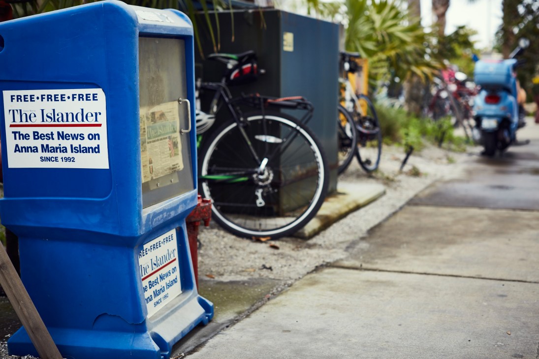 Blue newspaper dispensers for The Islander on a street with bicycles parked nearby.