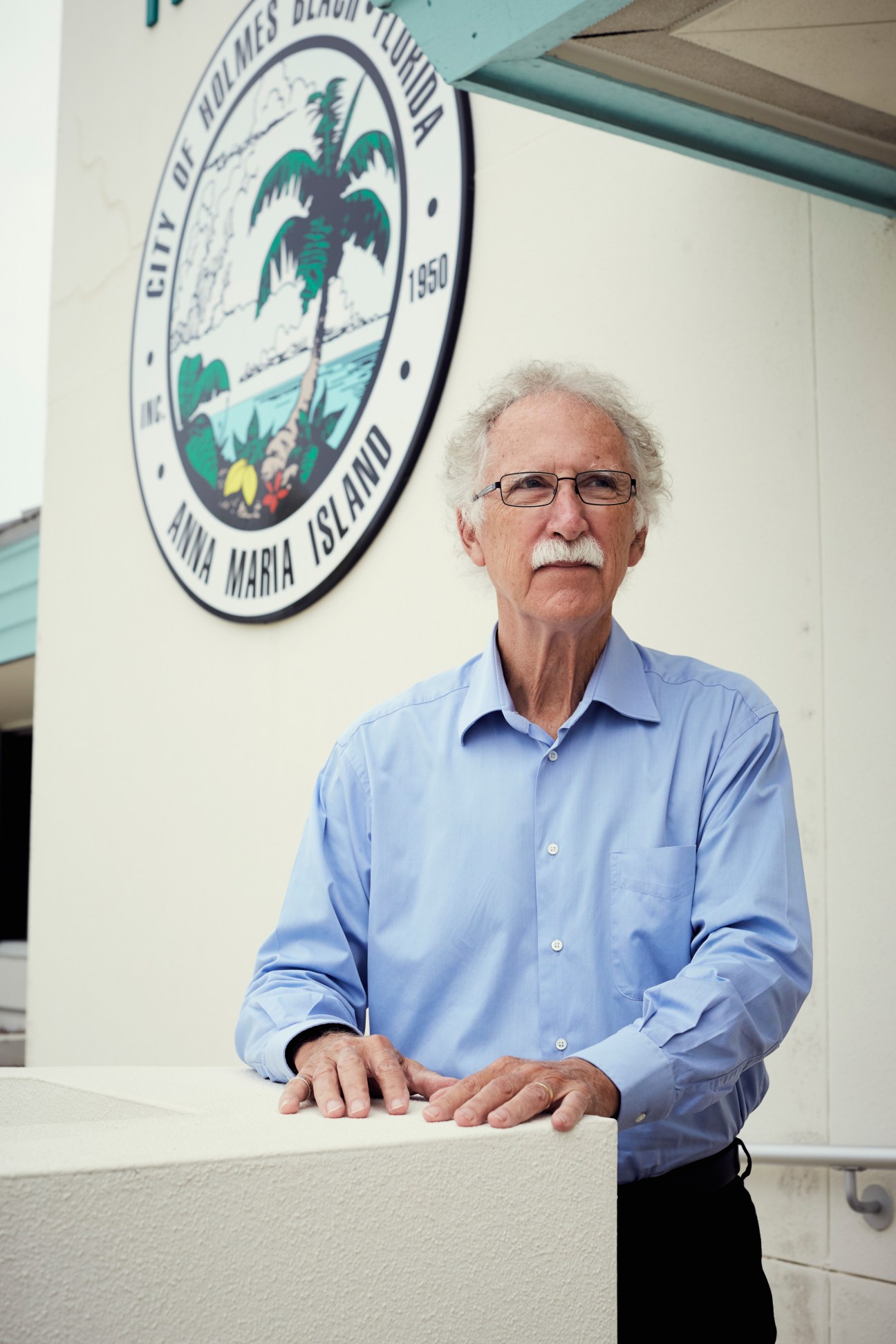 Holmes Beach mayor Bob Johnson stands in front of the Anna Maria Island city seal wearing a light blue shirt and glasses.