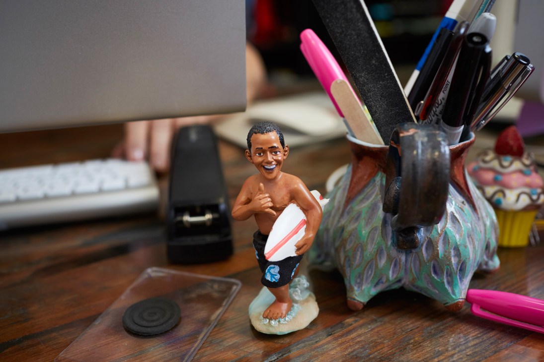 An Obama figurine holding a surfboard sits on a desk among office supplies and decorative items.