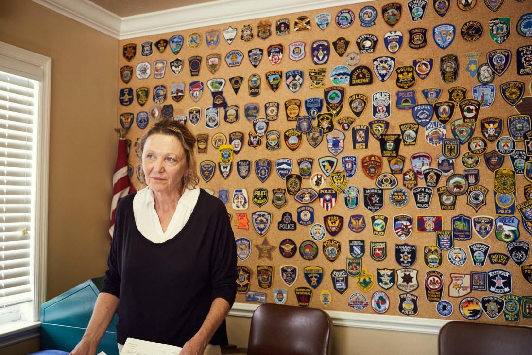 Reporter Kathy Prucnell stands in an office in front of a cork board displaying numerous police department badges and patches.