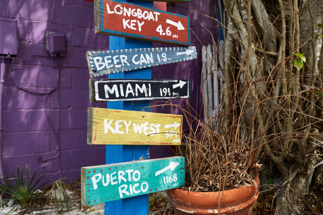 Colorful directional signs mounted on a pole against a purple wall, showing distances to various destinations including Longboat Key, Miami, Key West and Puerto Rico.