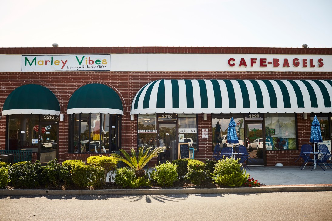 Storefront of The Islander newspaper office between Marley Vibes gift shop and Cafe-Bagels on a brick strip mall.