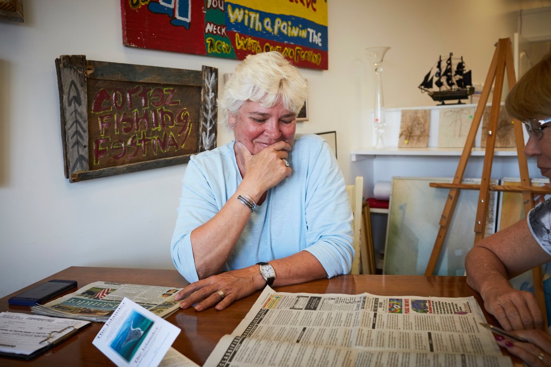 Woman with white hair in light blue shirt reads newspaper at desk in office.