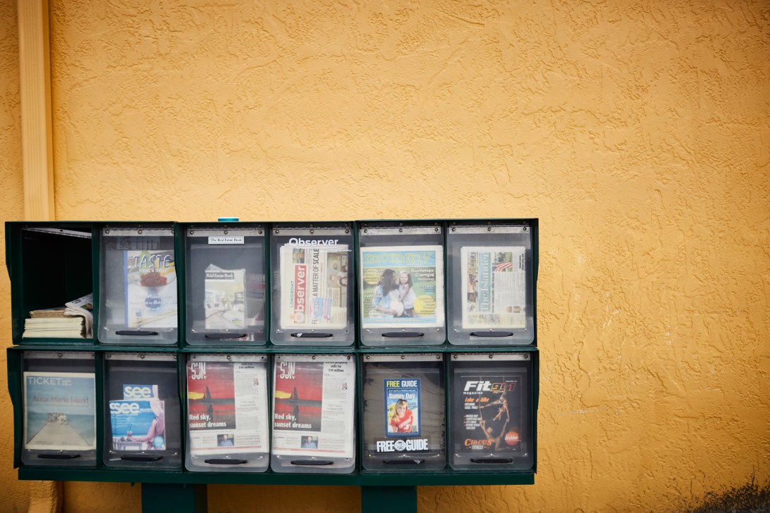 Newspaper and magazine vending boxes mounted on a green stand against a yellow stucco wall.