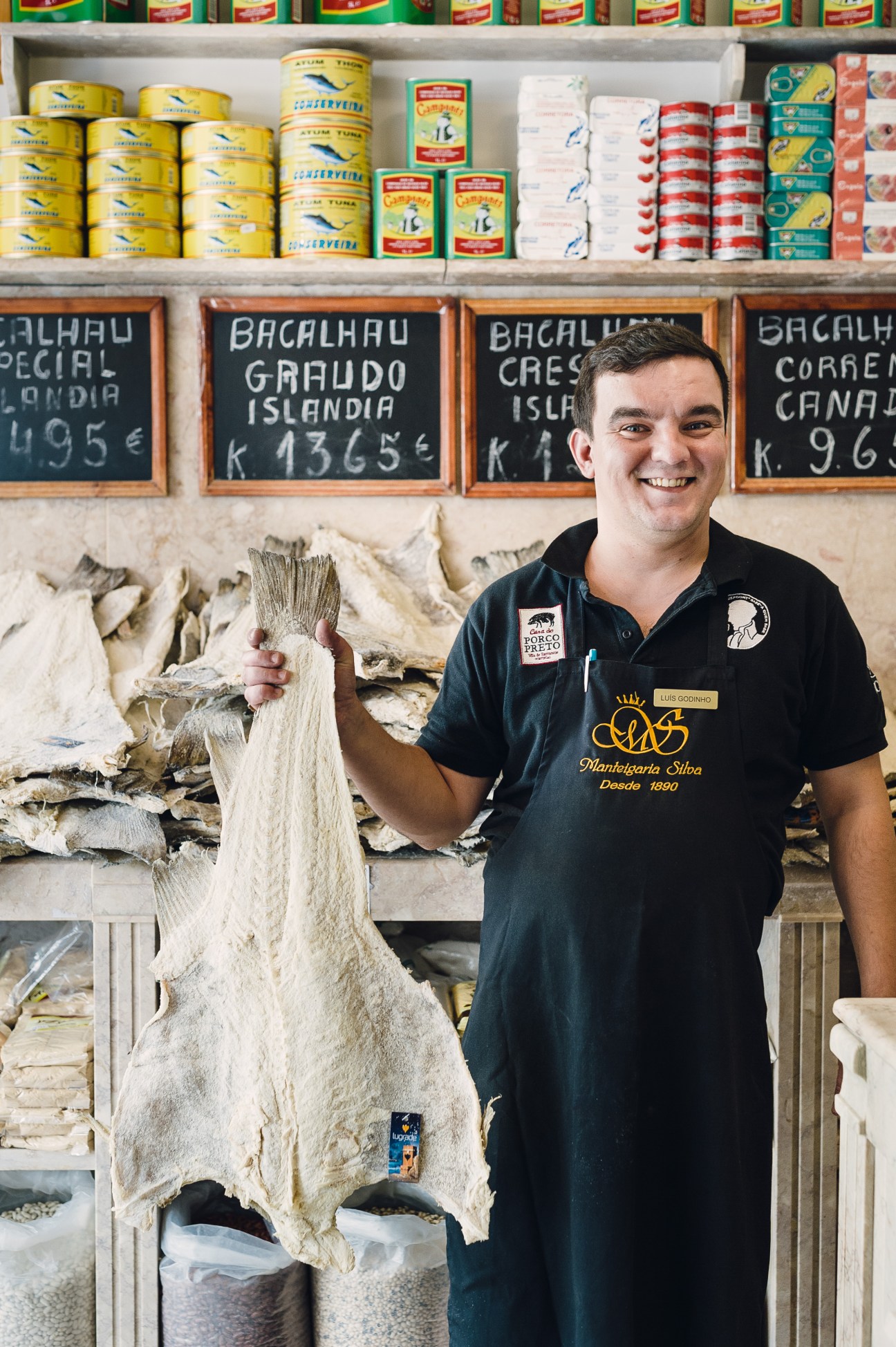 Man holding salt cod at Manteigaria Silva with shelves of tinned fish and products behind him in Lisbon.