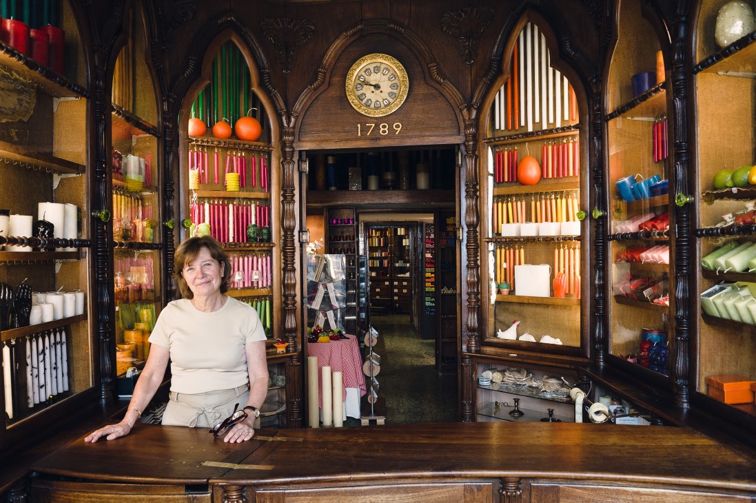 Woman standing behind counter in ornate wooden shop interior with 1789 clock and colorful candles displayed on shelves.