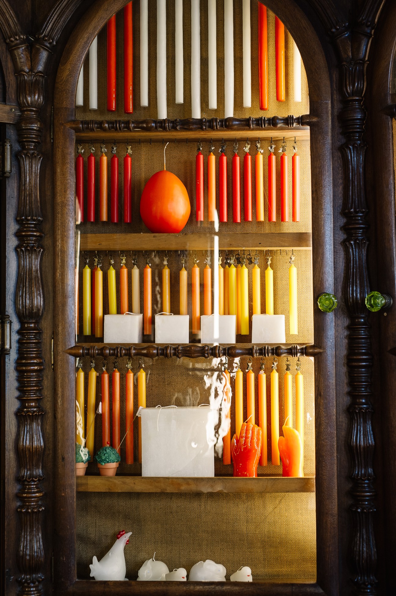 Display of colorful candles in red, orange and yellow arranged on shelves with white candle holders in an ornate wooden frame.