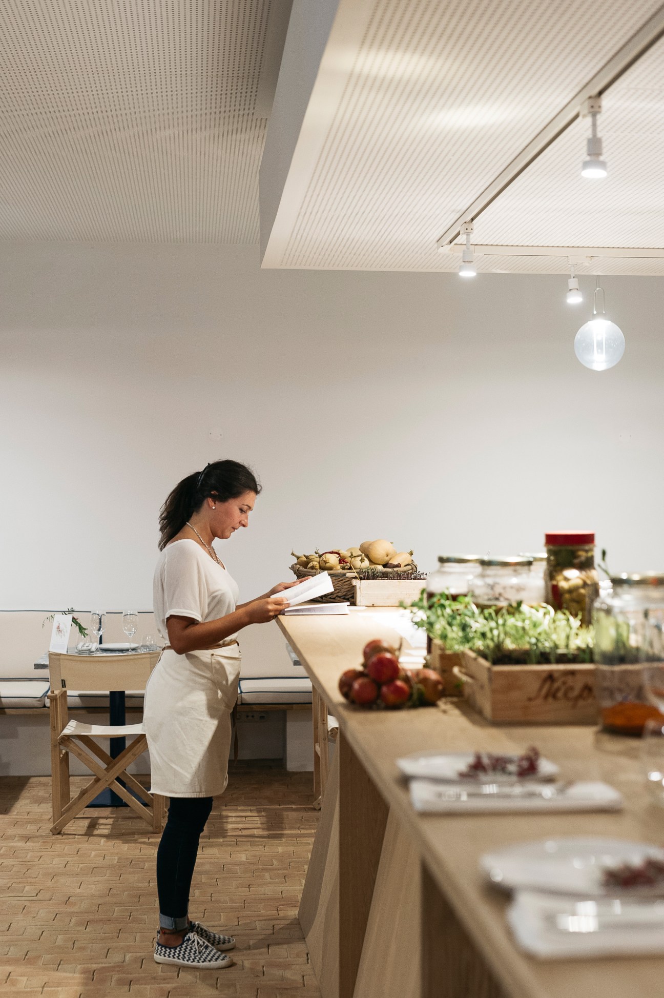 Woman in white apron reading from clipboard in bright kitchen with fresh produce displayed on counters.