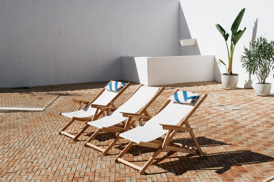 Three wooden deck chairs with white canvas and blue-striped pillows on a terracotta brick patio beside white walls and potted plants.