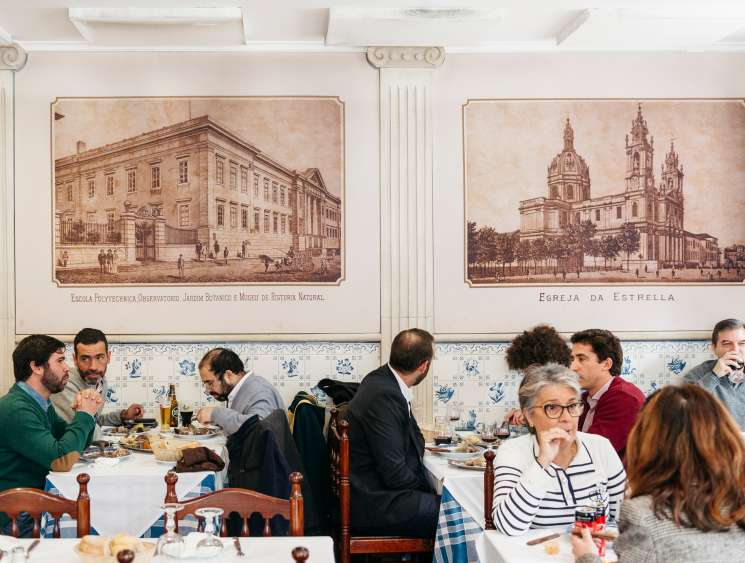 Diners eating at tables in Casa dos Pasarinhos restaurant with historic Lisbon paintings on white tiled walls
