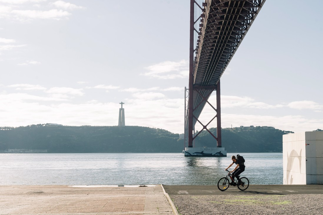 Cyclist riding on a waterfront cycle path beneath the 25 de Abril bridge in Lisbon with the Tagus river and monument visible.