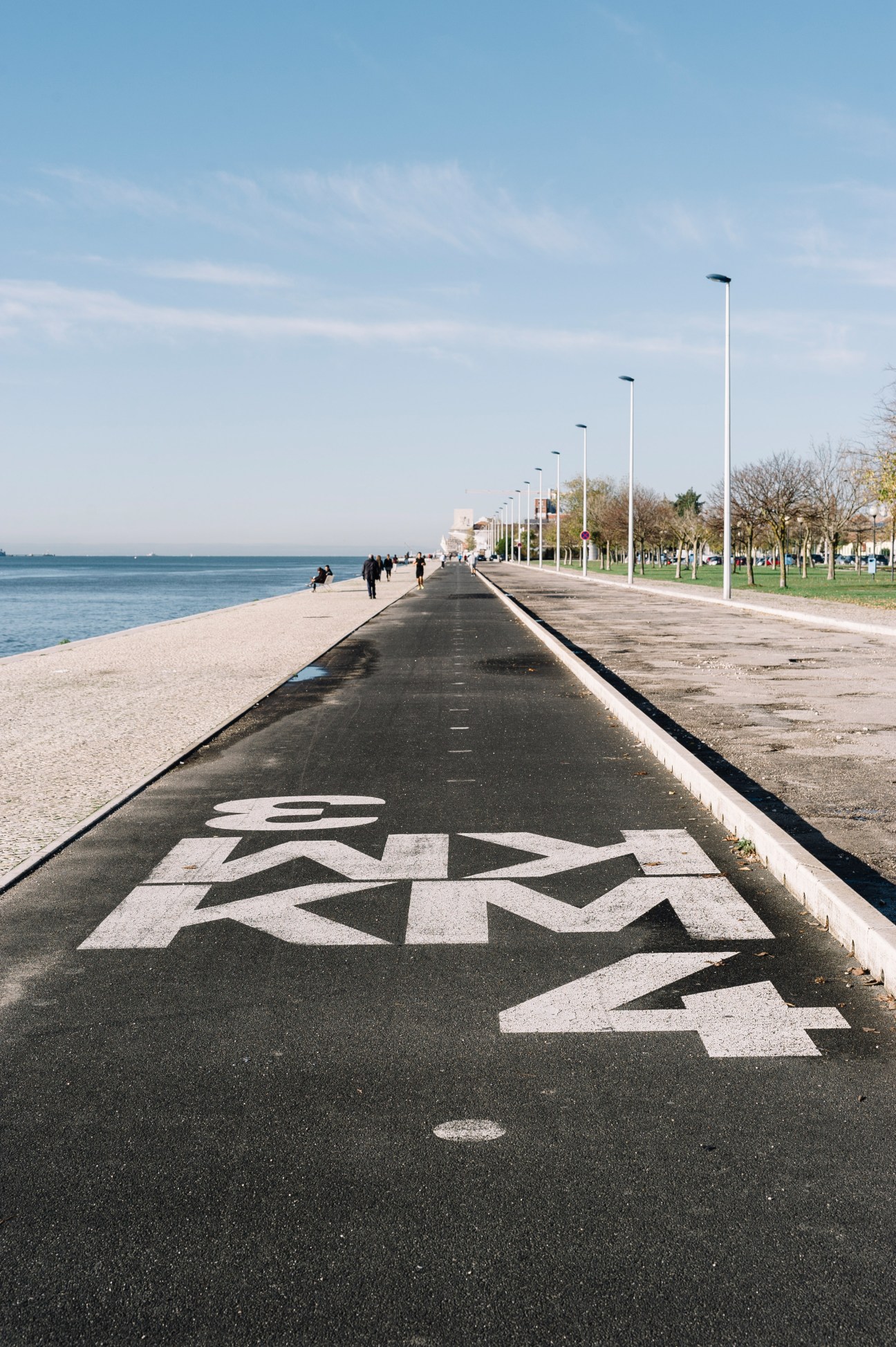 Waterfront cycle lane in Lisbon with white stencilled graphics on black asphalt and street lamps along the path.