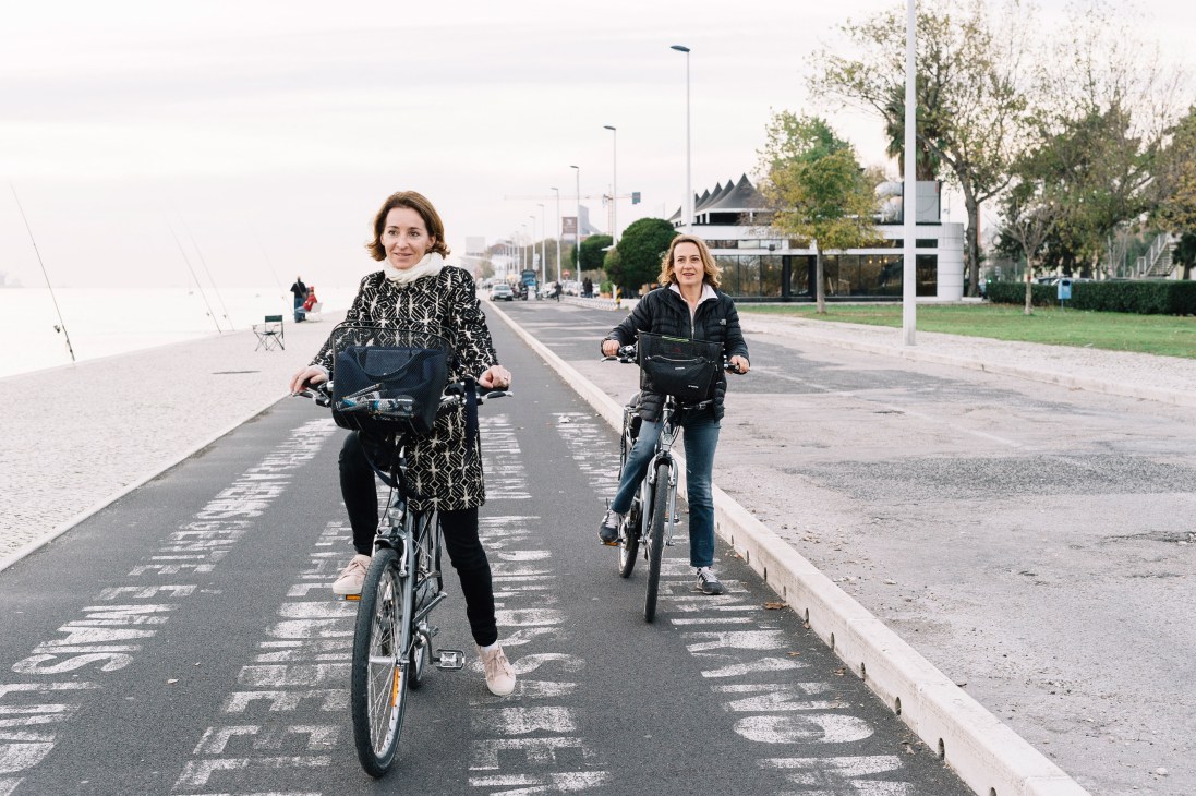 Two women cycling on a painted cycle lane beside Lisbon's waterfront near the Maat museum.